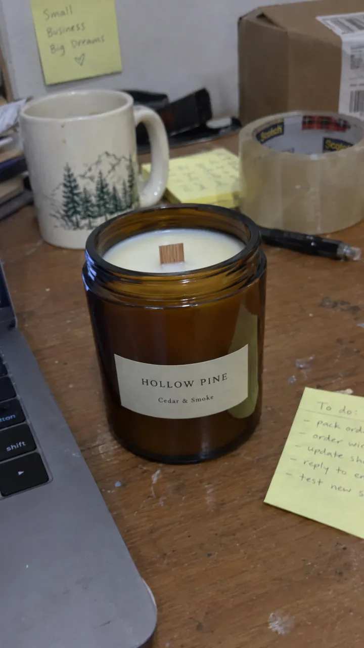 Phone photo of an amber jar candle on a cluttered desk with a mug, tape and sticky notes